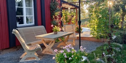 Trädgård med träbänkar, bord, blommor och röd husfasad i kvällssol.