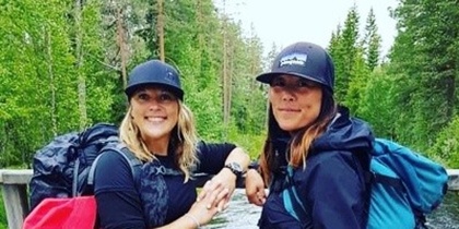Två kvinnor med ryggsäckar står på en träbro i skogen vid en forsande å.