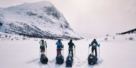 Fem personer med ryggsäckar och pulkor står på skidor i ett snötäckt fjällandskap med berg i bakgrunden.