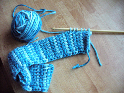 En virkad blå och vit halsduk med garnnystan och virknål på ett träbord.