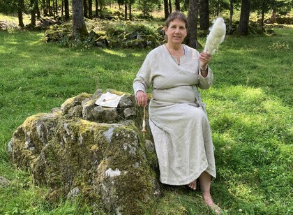 Kvinna i ljus klänning sitter på en mossig sten i skogen och spinner ull med slända.