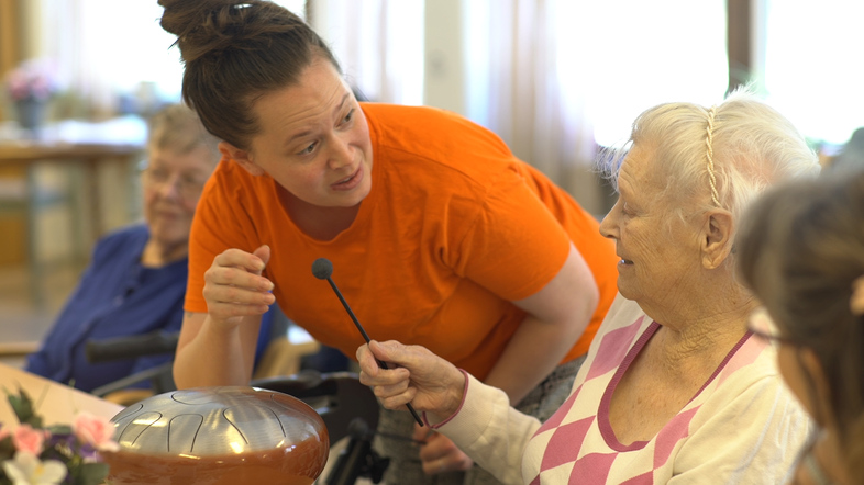 En yngre kvinna i orange tröja pratar med en äldre kvinna som håller i en mikrofon vid ett bord.