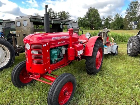 Röd veterantraktor står på en gräsmatta med andra fordon i bakgrunden under en molnig himmel.