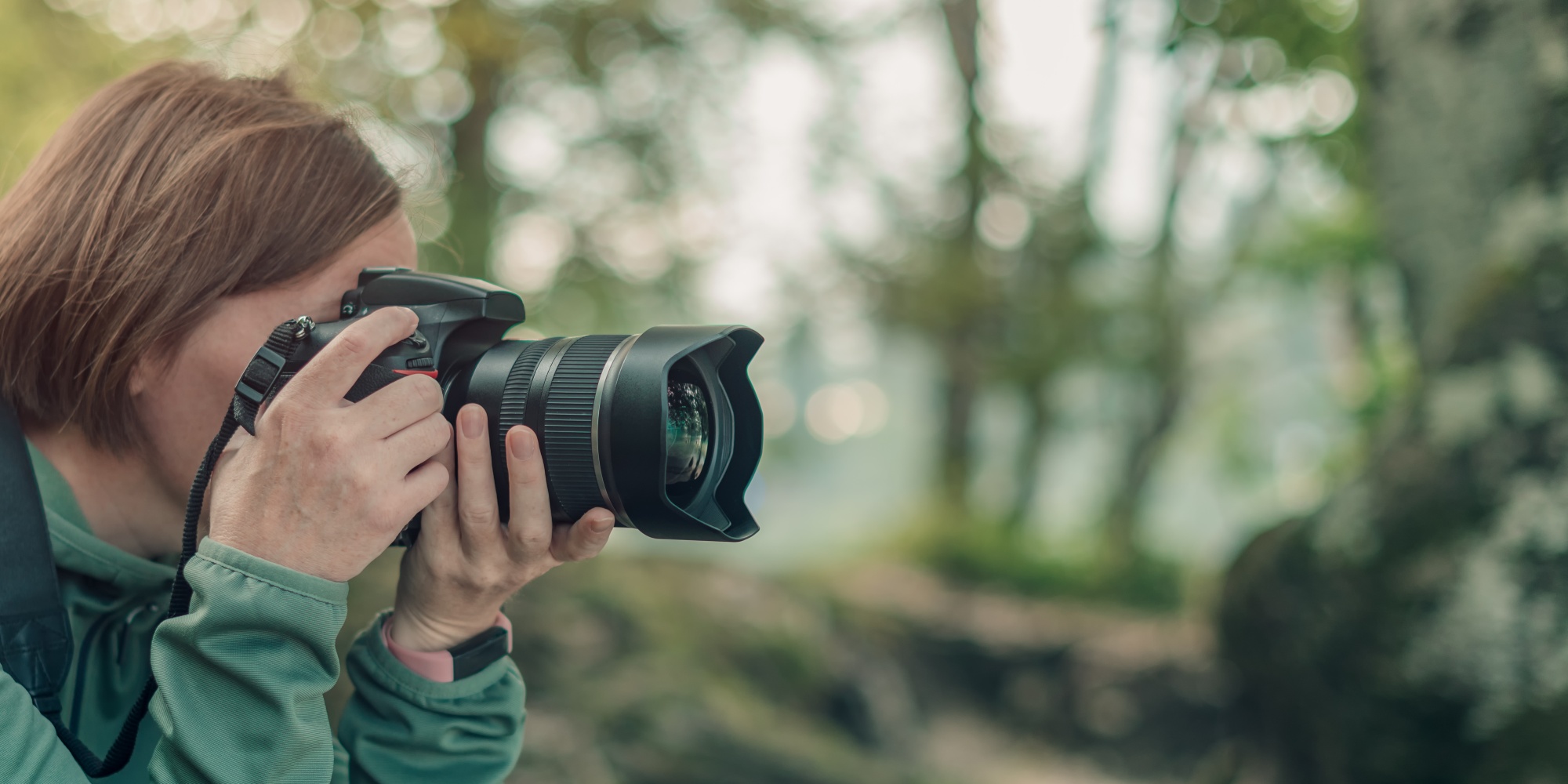 Person med kamera fotograferar i en skogsmiljö, klädd i grön jacka och ryggsäck.