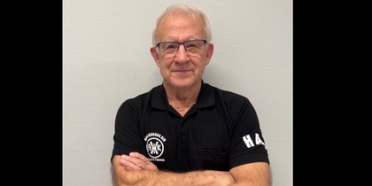 Man med armarna i kors, iförd svart pikétröja med vit text, står mot ljus vägg. Ansiktet är suddat.