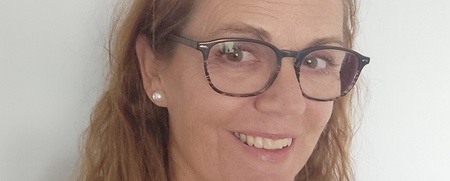 Kvinna med långt, vågigt hår och glasögon ler mot kameran framför en vit bakgrund.