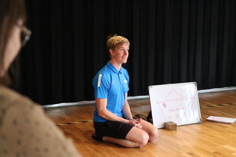 En person i blå tröja sitter på knä på ett trägolv framför en whiteboard med ritning, i ett rum med svarta gardiner.