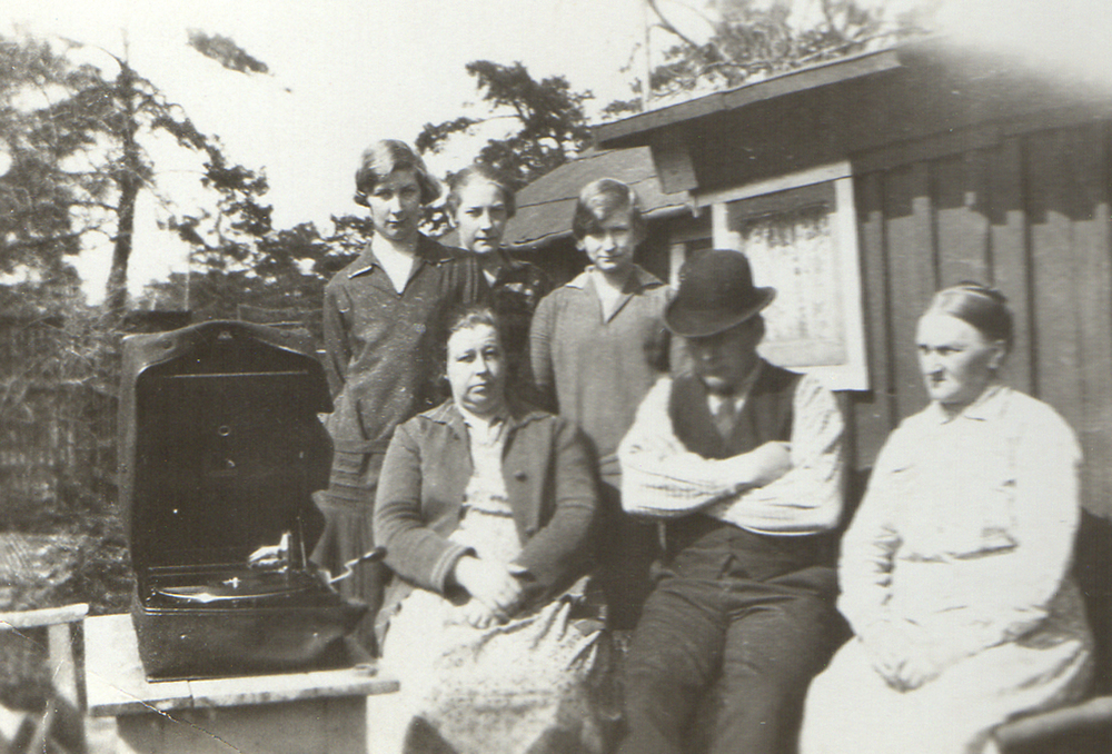 Gammalt svartvitt foto av fem personer utomhus vid ett hus, med en grammofon på ett bord.