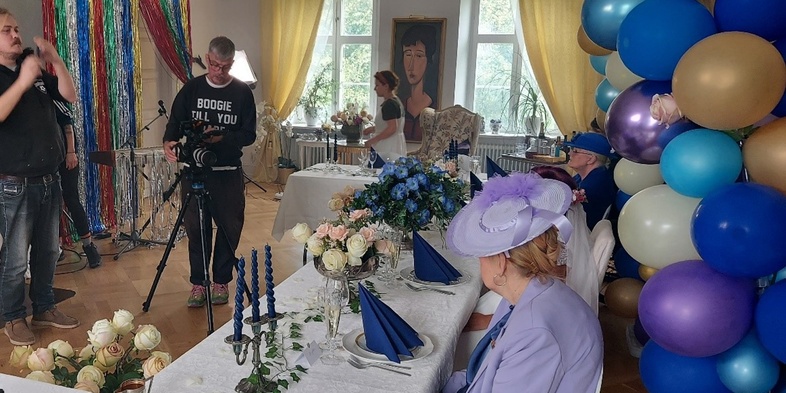 En bild av en filminspelning med en regissör, kameraman och två vackert klädda äldre damer som är skådespelare.