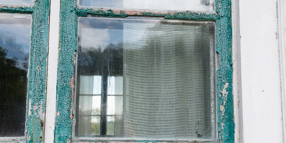 Närbild av ett gammalt fönster med avskavd grön färg och gardin på insidan.