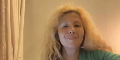 Kvinna med ljust lockigt hår sitter inomhus och spelar akustisk gitarr.