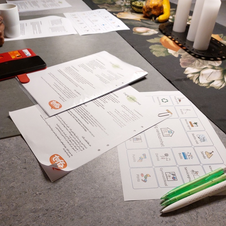 Ett bord med papper, pennor, mobil, kaffekoppar och dekorationer. Människor sitter runt bordet.