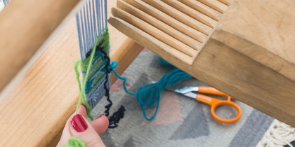 En person väver med garn på en vävstol, med händer och verktyg synliga.