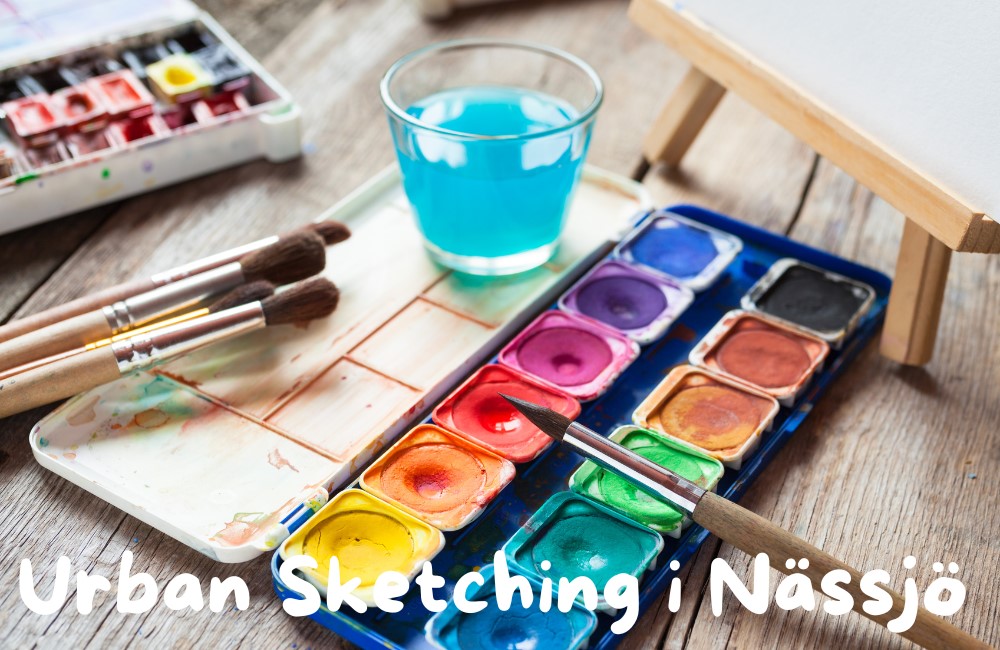 Akvarellfärger, penslar och ett glas vatten på ett träbord med texten Urban Sketching i Nässjö.