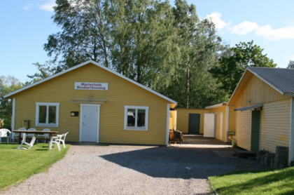 Gula trähus med vita dörrar och fönster, omgivna av grön gräsmatta och träd under blå himmel.