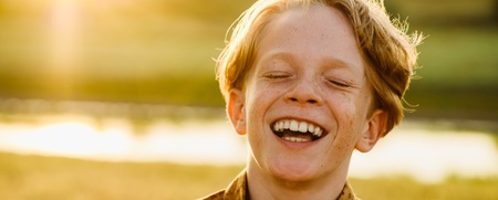 En ung pojke skrattar glatt utomhus i solnedgången med grönt landskap i bakgrunden.