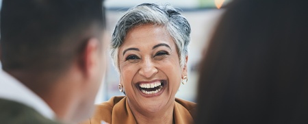 En leende kvinna med kort grått hår sitter vid ett bord och samtalar med två andra personer.