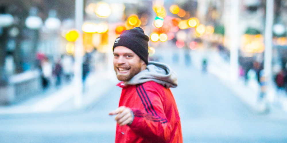 Person i röd jacka och svart mössa står på en stadsgata med suddig bakgrund och ljusprickar.