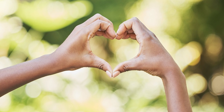 Två händer formar ett hjärta mot en suddig, grön bakgrund utomhus.