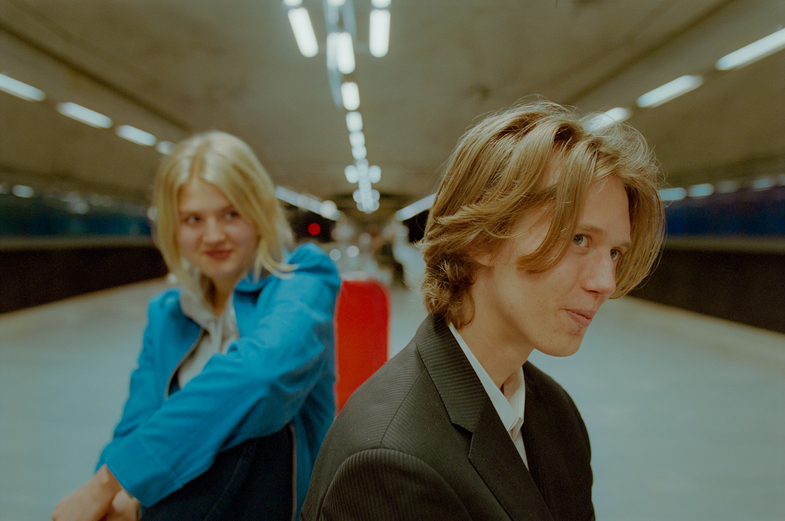 Två unga personer sitter på en tunnelbaneperrong, en i förgrunden och en i bakgrunden, båda tittar åt olika håll.