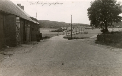 Svartvit bild av en landsväg med byggnader till vänster och ett träd till höger, landskap i bakgrunden.