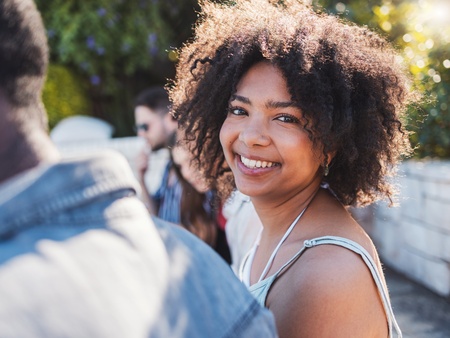Ung kvinna med lockigt hår ler mot kameran utomhus i solskenet, med andra personer i bakgrunden.