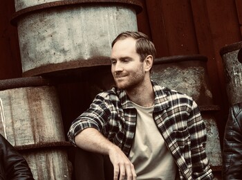 Tre personer sitter avslappnat framför staplade oljefat mot en trävägg, klädda i läderjackor och hattar.