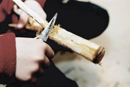 En person täljer en träpinne med en kniv, närbild på händer och trä.