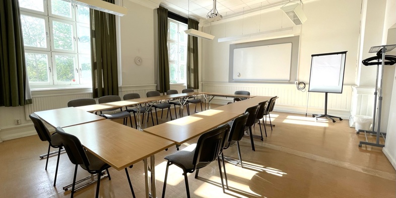 Ljust konferensrum med U-formade bord, stolar, stora fönster, whiteboard och blädderblock.