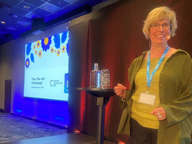 Kvinna står på scen bredvid ett bord med vattenflaska och glas, presentation visas på stor skärm.