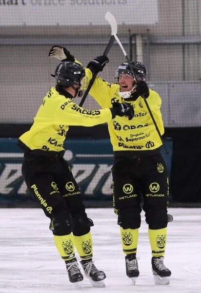 Två bandyspelare i gula och svarta dräkter firar på isen under en match.