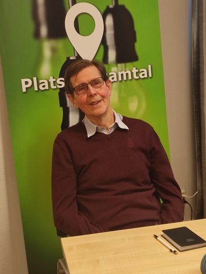 Man med glasögon sitter vid ett bord framför en grön skylt med texten 'Plats för samtal'.