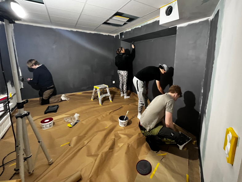 Fyra personer målar väggarna i ett rum med mörk färg, golvet täckt med skyddspapper.