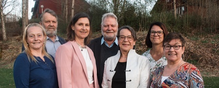 Sju vuxna personer står utomhus i grupp och ler mot kameran, med träd och hus i bakgrunden.