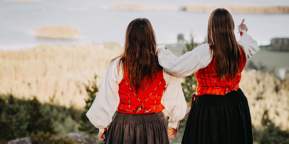 Två personer i traditionella folkdräkter står på en klippa och ser ut över ett landskap med vatten och skog.