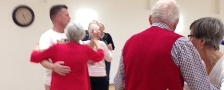 Flera äldre par dansar tillsammans i en ljus sal med trägolv och en klocka på väggen.