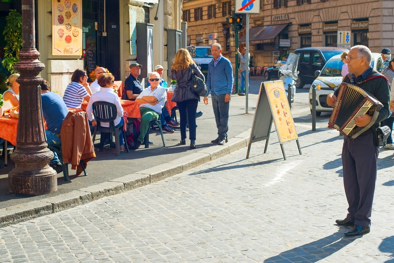 En gatumusikant spelar dragspel framför ett café med uteservering på en solig stadsgata.