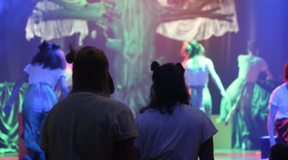 Personer i kostym sitter och tittar på en teaterföreställning med färgglad belysning och en stor trädrekvisita på scenen.