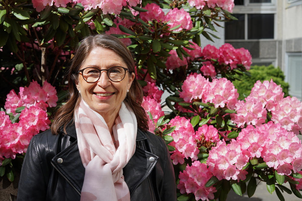 Leende kvinna med glasögon och ljus sjal står framför rosa blommande buskar utomhus.