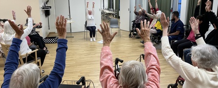 En grupp äldre personer sitter i en cirkel och gör sittande gymnastik med armarna uppsträckta.