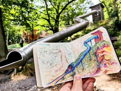 En hand håller en skissbok med en färgglad teckning av en rutschkana, framför den riktiga rutschkanan i en park.