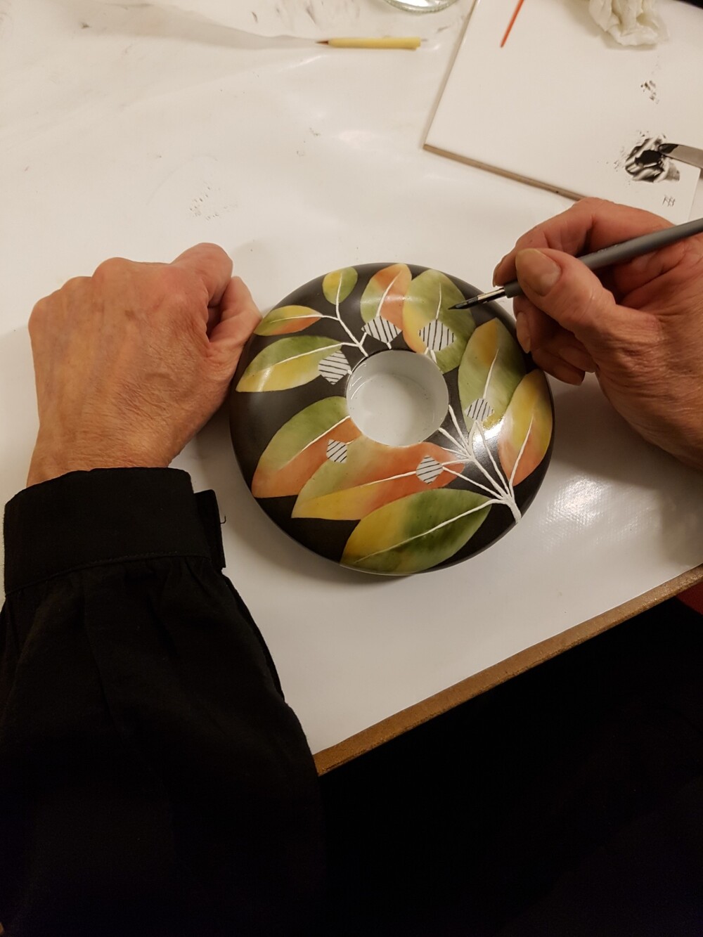 En person målar ett keramiskt fat med bladmotiv i grönt, gult och orange på en vit arbetsyta.