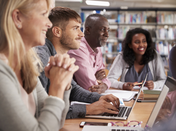 Fyra vuxna personer sitter vid ett bord i ett bibliotek och arbetar tillsammans med datorer och anteckningar.