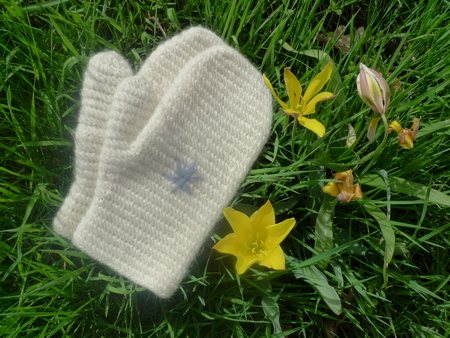 Ett par vita stickade vantar ligger på gräs bredvid gula blommor.