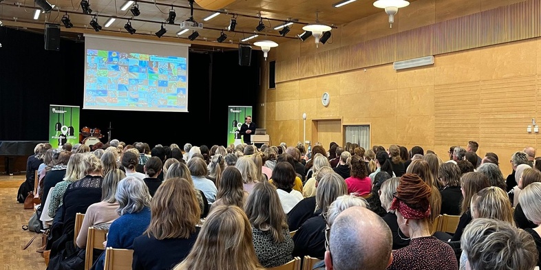 En stor grupp människor sitter i en aula och lyssnar på en presentation med bildspel på en scen.