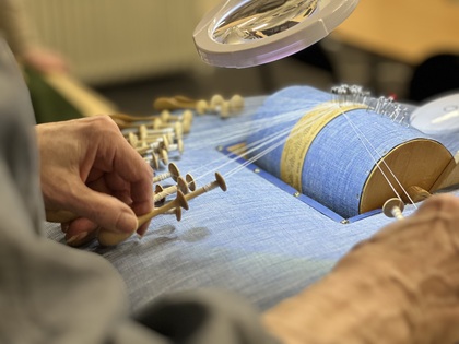 Närbild av händer som tillverkar spets med hjälp av knyppelpinnar och en blå knyppeldyna.