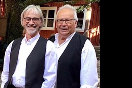 Två personer i vita skjortor och svarta västar står utomhus på en stenlagd gård med träbyggnad i bakgrunden.