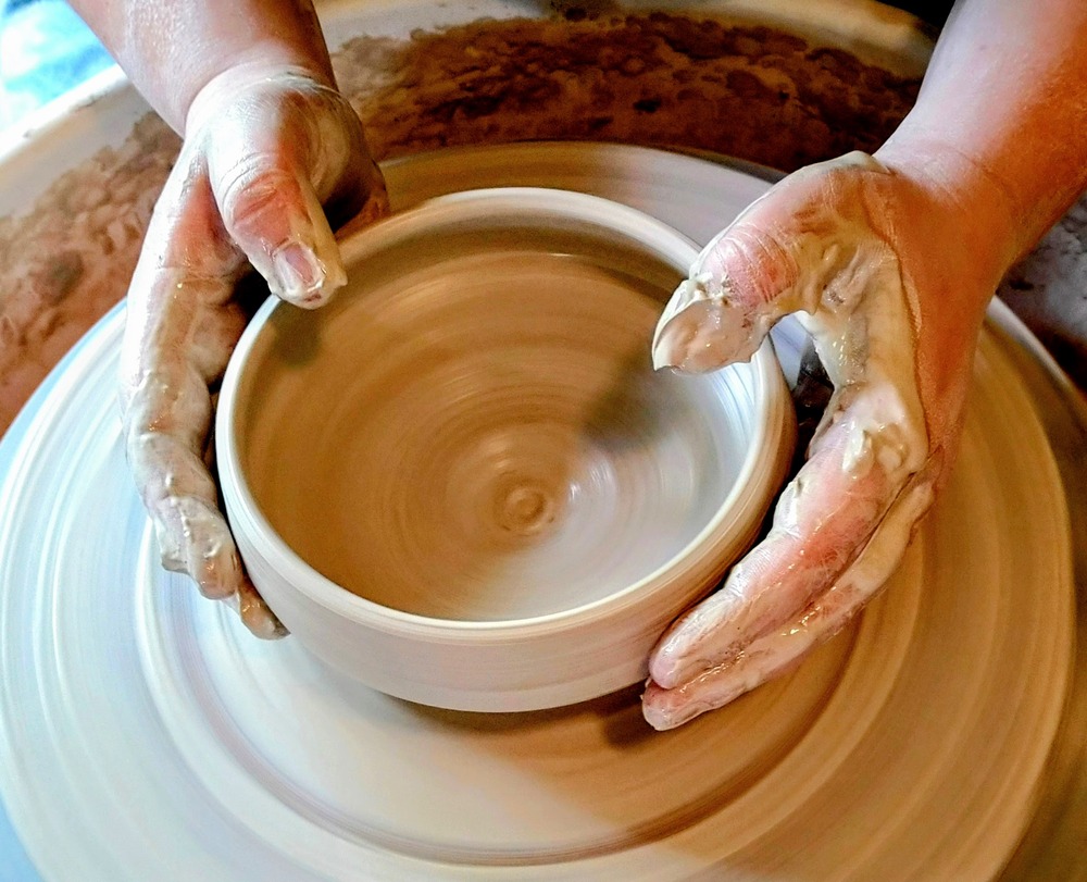 Två händer formar en lerkruka på en drejskiva. Händerna är täckta av lera.