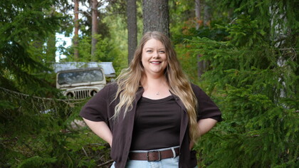 Leende kvinna med långt blont hår står i en grön skog, med en gammal bil i bakgrunden.