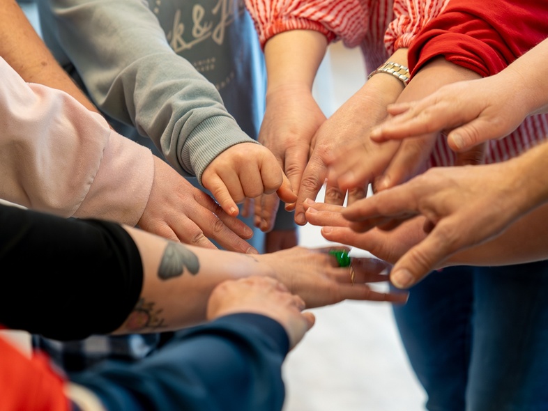 Flera personer sträcker in sina händer mot mitten i en gemensam cirkel, fokus på händerna.